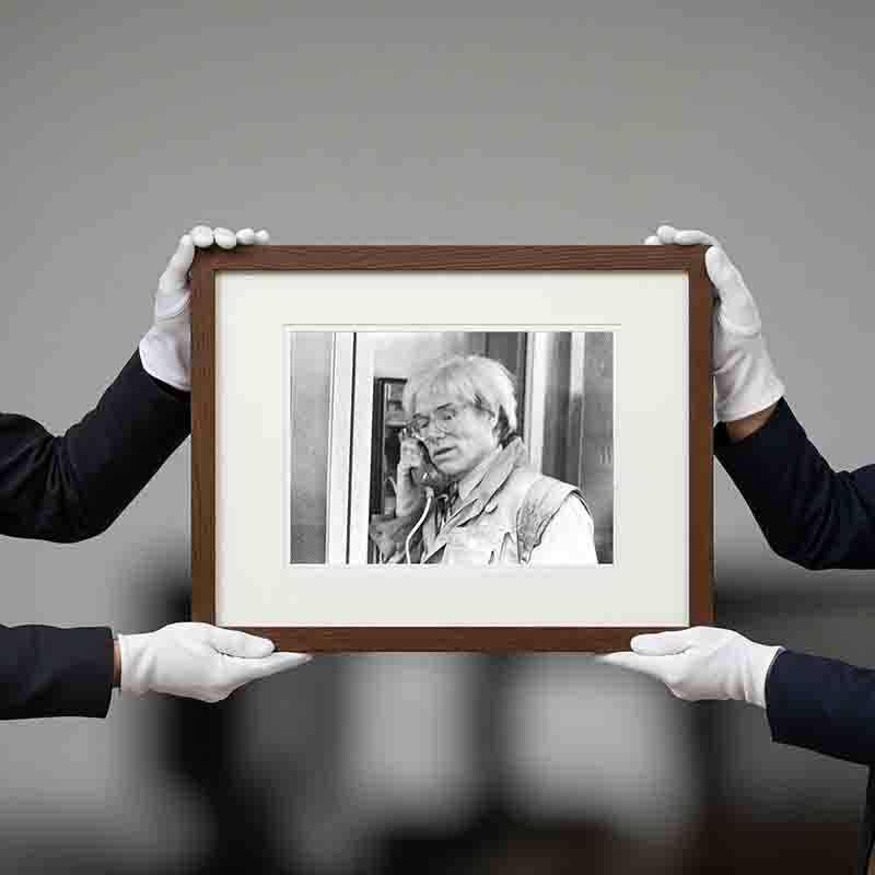 Two people, wearing black shirts and white gloves, are holding the framed black and white Bock-Schroeder photograph of Andy Warhol at an auction. The photograph, which is a close-up of Warhol with his famous white hair and glasses, shows him holding a phone receiver to his ear with a thoughtful expression.