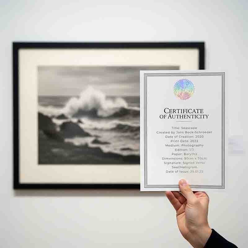 A close-up image showing a framed black-and-white photograph of a dramatic seascape with crashing waves, hanging on a white gallery wall. In the foreground, a person's hand, wearing a dark sleeve, holds up a 'Certificate of Authenticity' card.