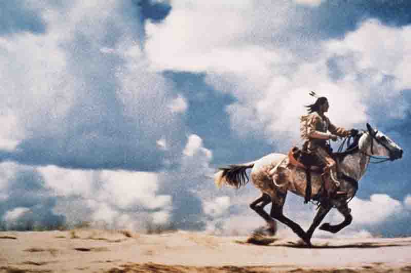 A full-shot image shows a person with long, dark hair wearing a headdress with a single feather, riding a white horse at a gallop across a sandy, barren landscape. The person is in profile, facing toward the right side of the image. The horse is kicking up a cloud of dust, and the sky above is filled with dramatic white and gray clouds.