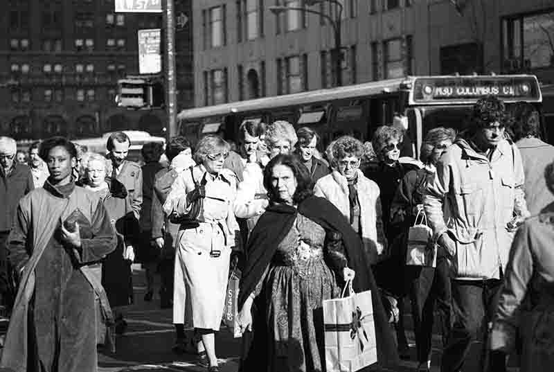 A black and white street photograph of a crowded sidewalk in New York City. In the foreground, a woman in a dark cape looks toward the camera while holding a shopping bag. Behind her, a diverse crowd of people is walking, and a city bus with a sign for B'WAY & COLUMBUS CIR is visible in the background.