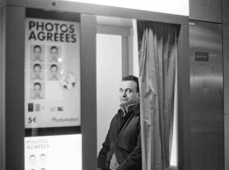 Black and white photograph depicting a man partially visible inside a photo booth, looking directly towards the viewer with a slightly intense or curious expression.