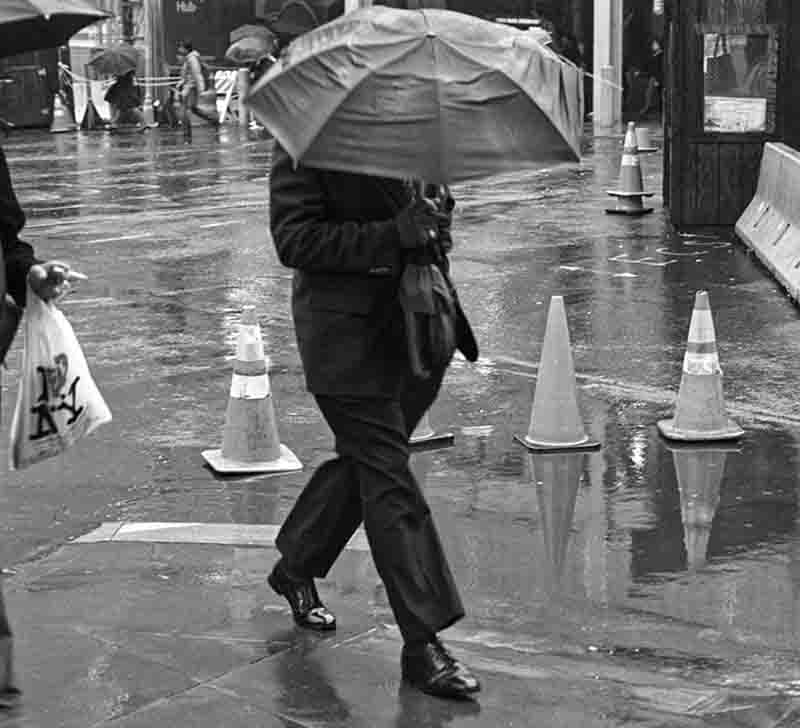 A black and white photograph of a person walking on rain-slicked Ground Zero in New York City, holding an umbrella that obscures their head. The wet pavement reflects the light from street lamps and the shapes of several traffic cones in the background. 