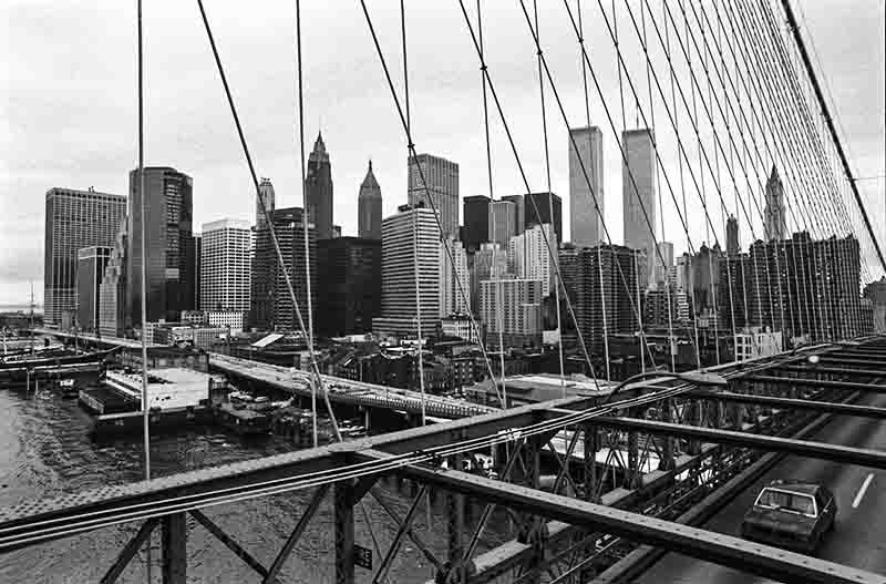 A black and white panoramic view of the lower Manhattan skyline as seen from the Brooklyn Bridge. The bridge's intricate network of cables and girders dominates the foreground, with a vehicle visible on the roadway. In the background, the dense cityscape includes the distinctive twin towers of the World Trade Center, surrounded by other skyscrapers.