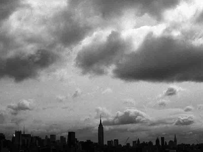A black and white photograph of the New York City skyline under a cloudy sky. The Empire State Building is prominently featured in the center, towering over other skyscrapers.