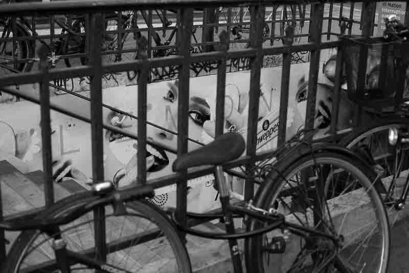 Black and white photograph of bicycles parked against a metal railing overlooking a stylized Schweppes advertisement billboard featuring graphic illustrations of eyes and lips.