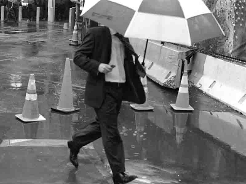 A well-dressed man in a suit and tie strides through the rain, holding an umbrella to shield himself from the downpour.