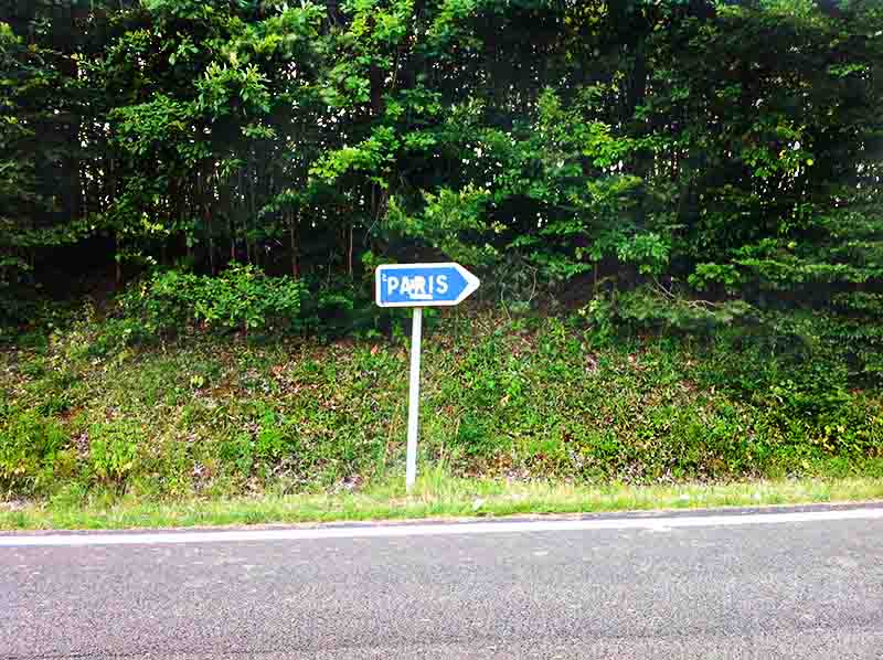 The photo shows a blue road sign pointing to the right, with the word PARIS written in white capital letters inside a white rectangle. The sign is mounted on a white pole.