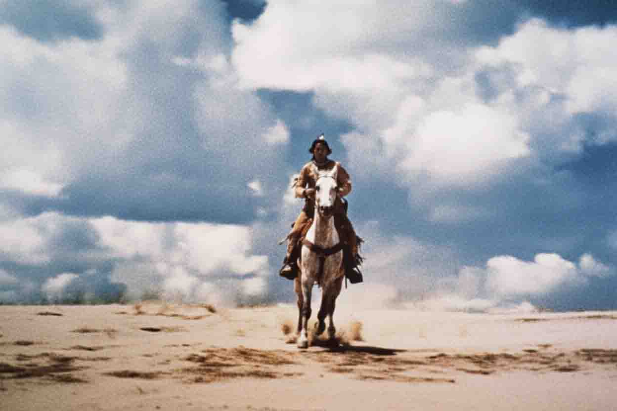 A person on horseback is shown riding toward the camera on a sandy, barren plain. The rider is wearing a headdress with what appears to be a single feather. The horse is light-colored and seems to be kicking up dust as it moves. The sky behind the rider is filled with large, dramatic white and gray clouds.