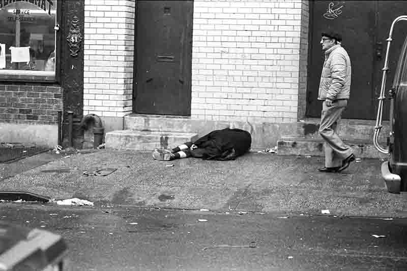 A black and white photograph of an urban street scene in New York City. In the foreground, a person is lying face down on the pavement next to the base of a brick building. In the background, another person is walking by, facing away from the camera. 