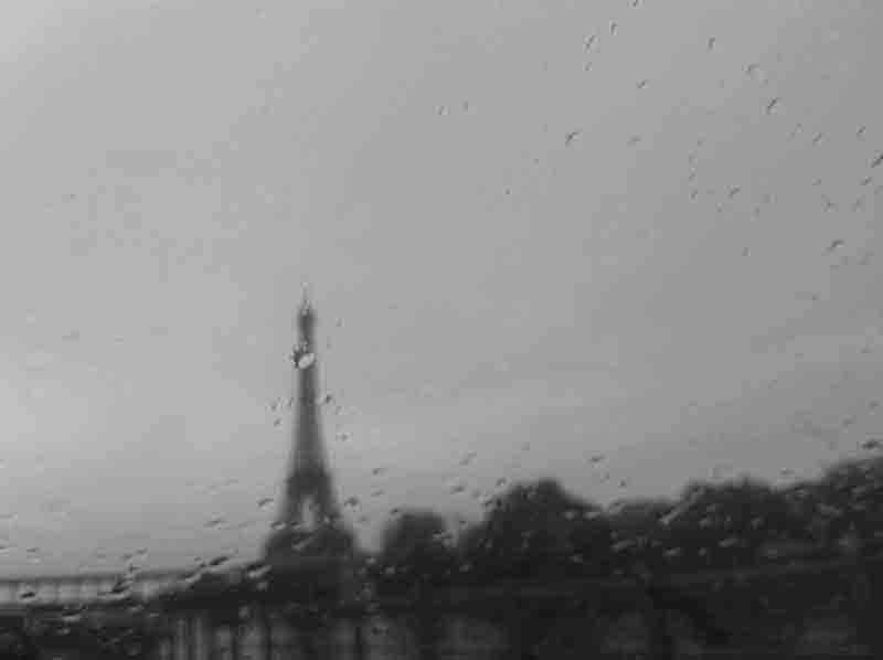 The Eiffel Tower is visible through a rain-soaked window, creating a blurred and atmospheric view of the iconic landmark.