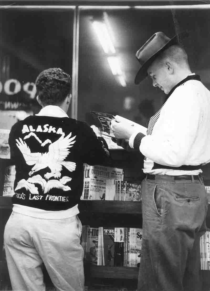 Two men stand outside a bookstore, engaged in reading, with shelves of books visible in the background.