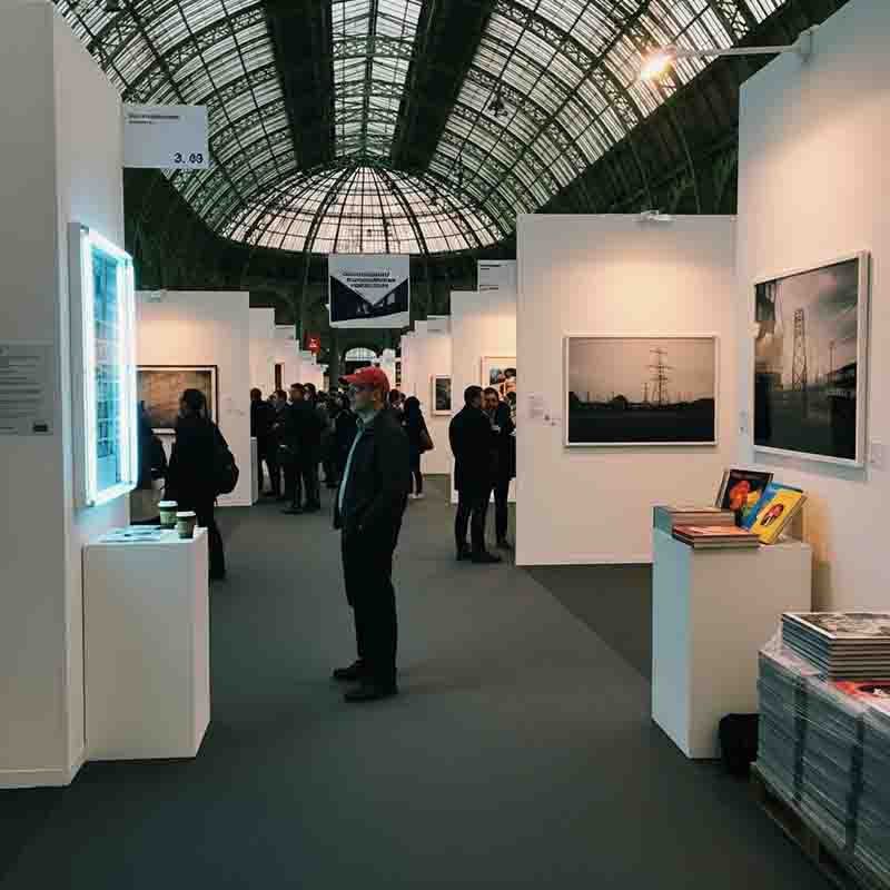 The image shows the interior of the Grand Palais in Paris, during the Paris Photo fair. The most prominent feature is the building's stunning architecture, particularly its immense glass roof and domed nave, which allows natural light to fill the vast space.